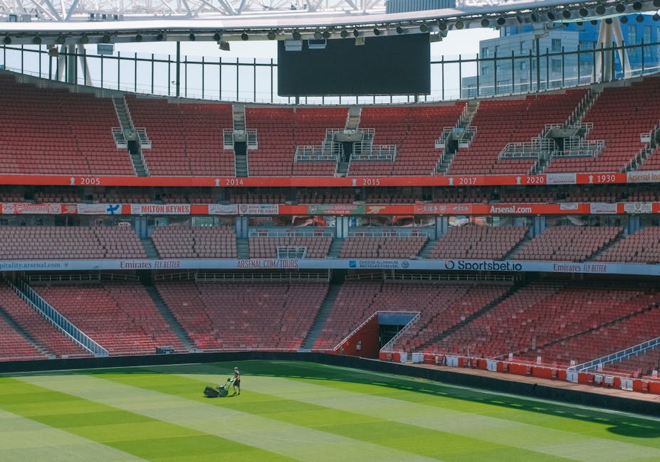 A view of the interior seating area of Tottenham Hotspur Stadium, featuring multiple rows of blue and red stadium seats arranged in tiers. The seats are mostly unoccupied, with a few scattered spectat