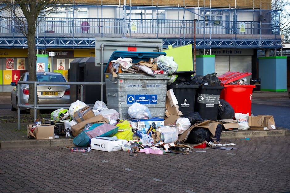 Two large black rubbish bags and a cardboard box filled with assorted waste rest on the pavement beside a black metal fence with vertical bars. The waste bags appear to contain household or garden ref